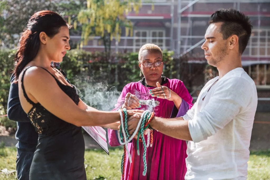 An officiant dressed in a purple toga has woven a smudging unity ceremony into the ceremony. A man wearing a white shirt and trousers, and a woman wearing a black laced dress are looking at the smudge stick.