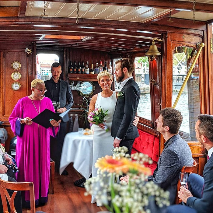  black marriage officiant dressed in purple gown, bride in white wedding dress and groom in navy blue suit on a Dutch boat