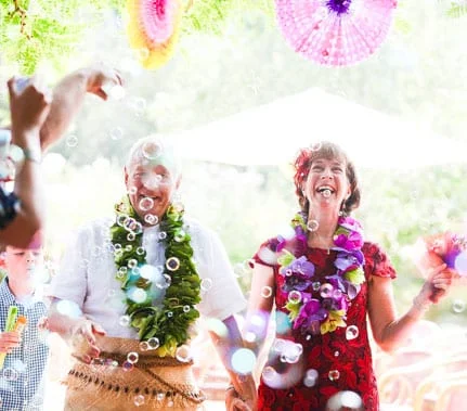 An older couple is getting married. The groom wears a white shirt and Hawaiian wedding clothes, and the bride wears a red dress. They stand together under a bubble of confetti as they leave their garden wedding in Amersfoort, the Netherlands.