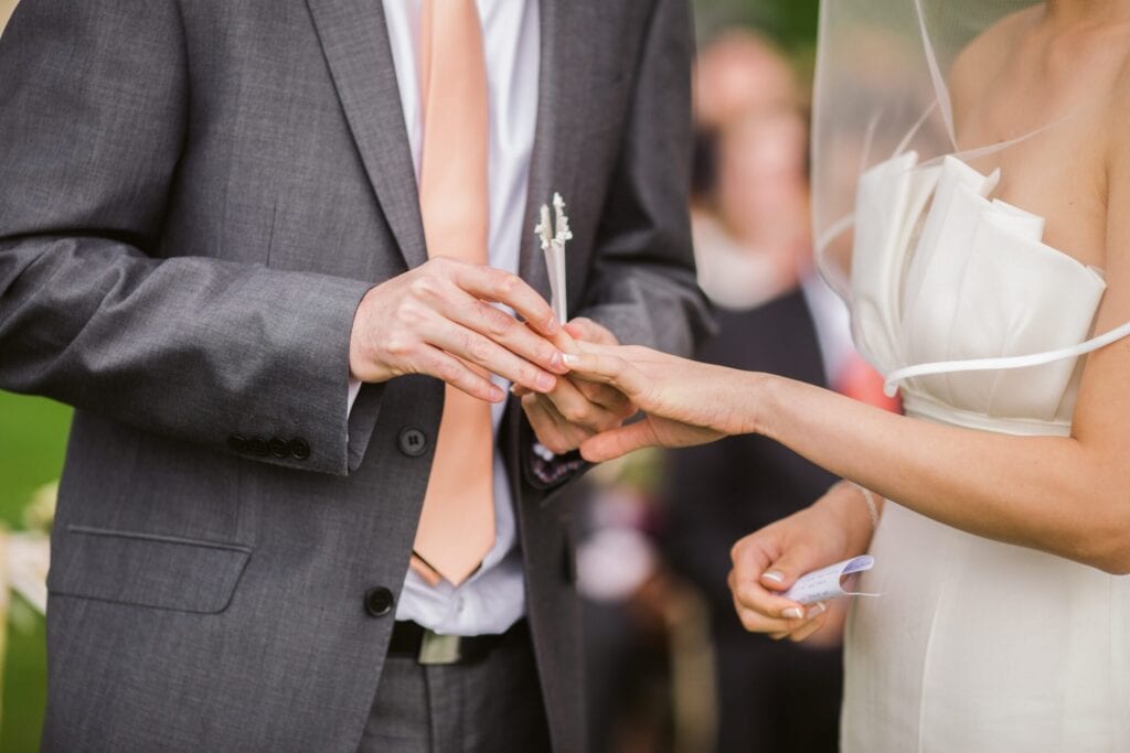 huwelijksrituelen groom in grey suit with orange tie putting wedding ring on his bride dressed in white satin dress with veil