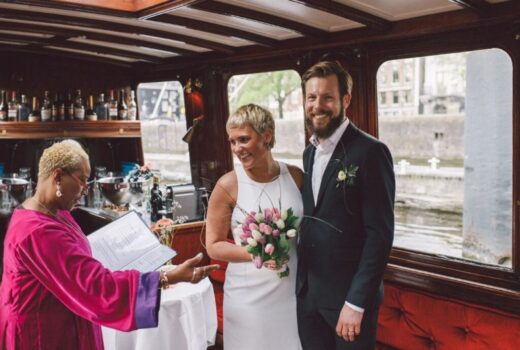 Getting Married Aboard, a an Amsterdam canal boat woman wearing a white bridal dress holding a pink and white floral bouquet and the groom smiling wearing a dark suit without tie.
