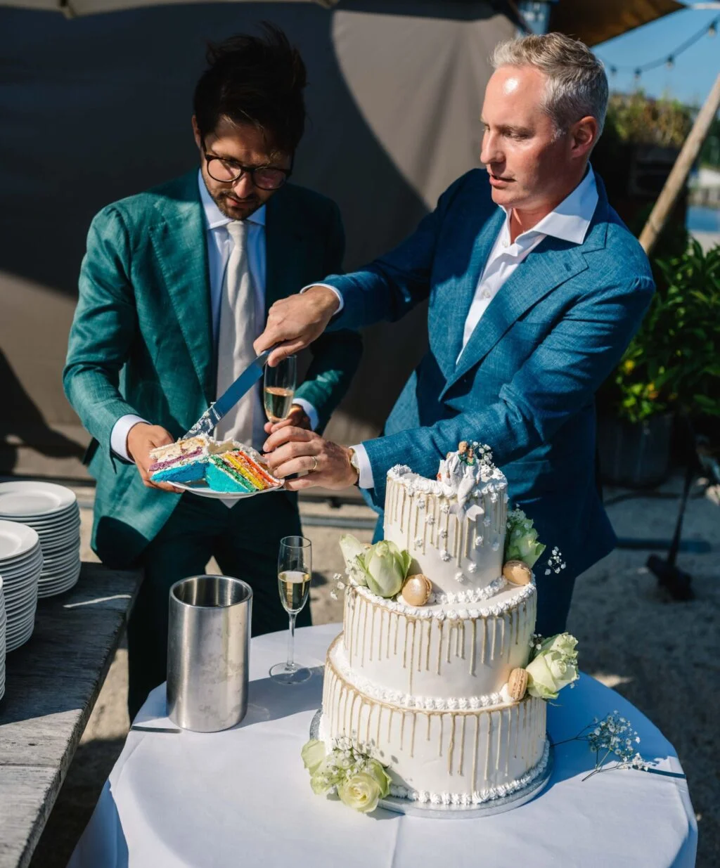 Ceremony rituals rite of passage woven into the ceremony. Gay couple cutting a rainbow wedding cake together. Both grooms are dressed in sky blue tweed and satin tuxedo suits.
