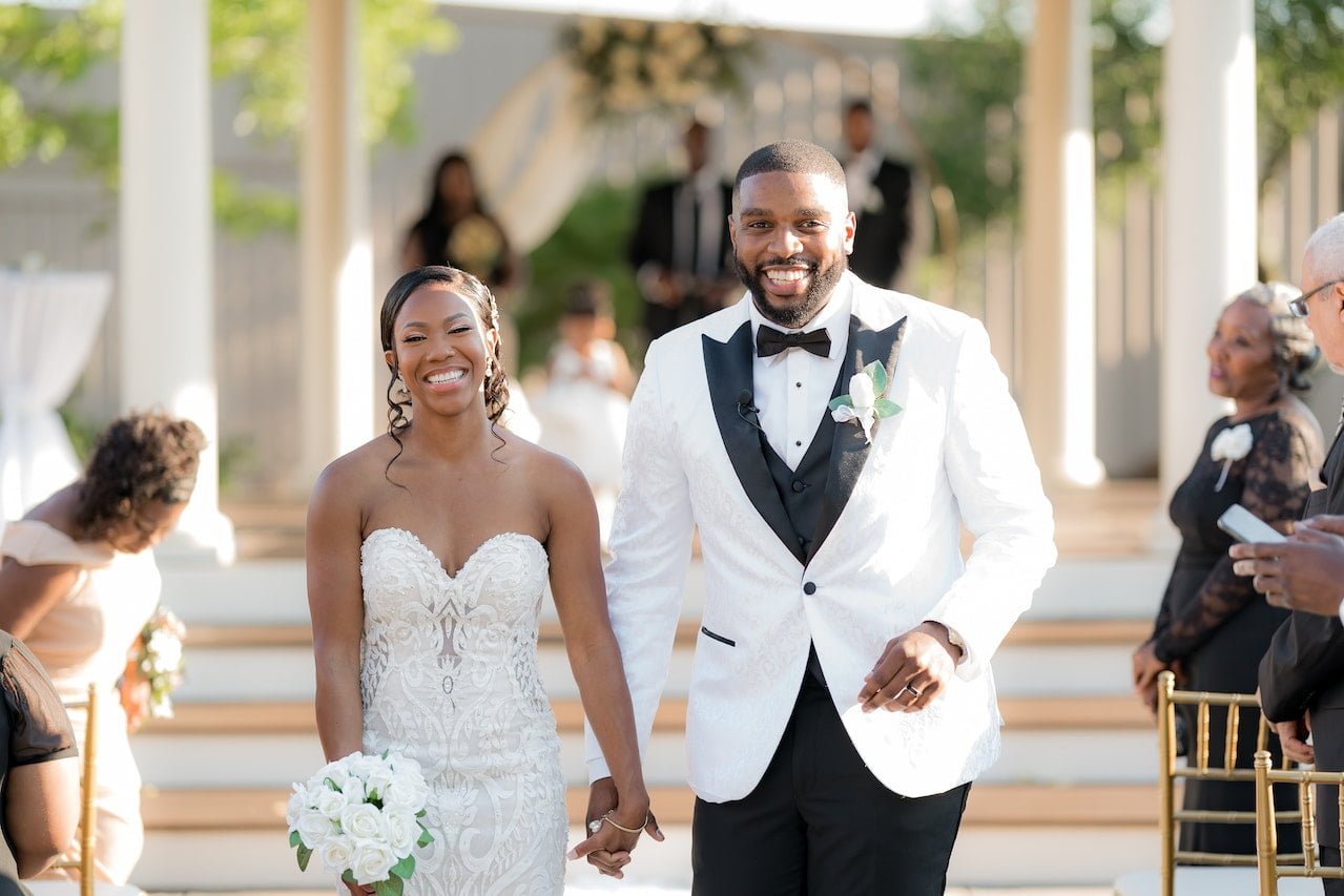 A luxurious wedding venue where a happy bride and groom walk together, smiling hand in hand, as the officiant and guests watch them leave the ceremony.