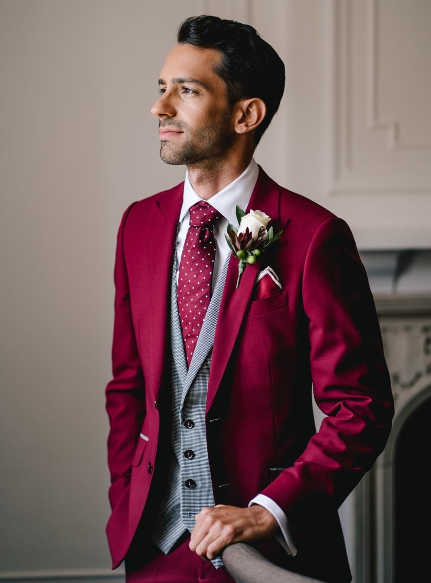 Waarom heb je een conflict over het organiseren van je bruiloft? bruiloft outfits en een bruidegom in een burgundy trouwpak en grijze gilet met stropdas staat te lachen.