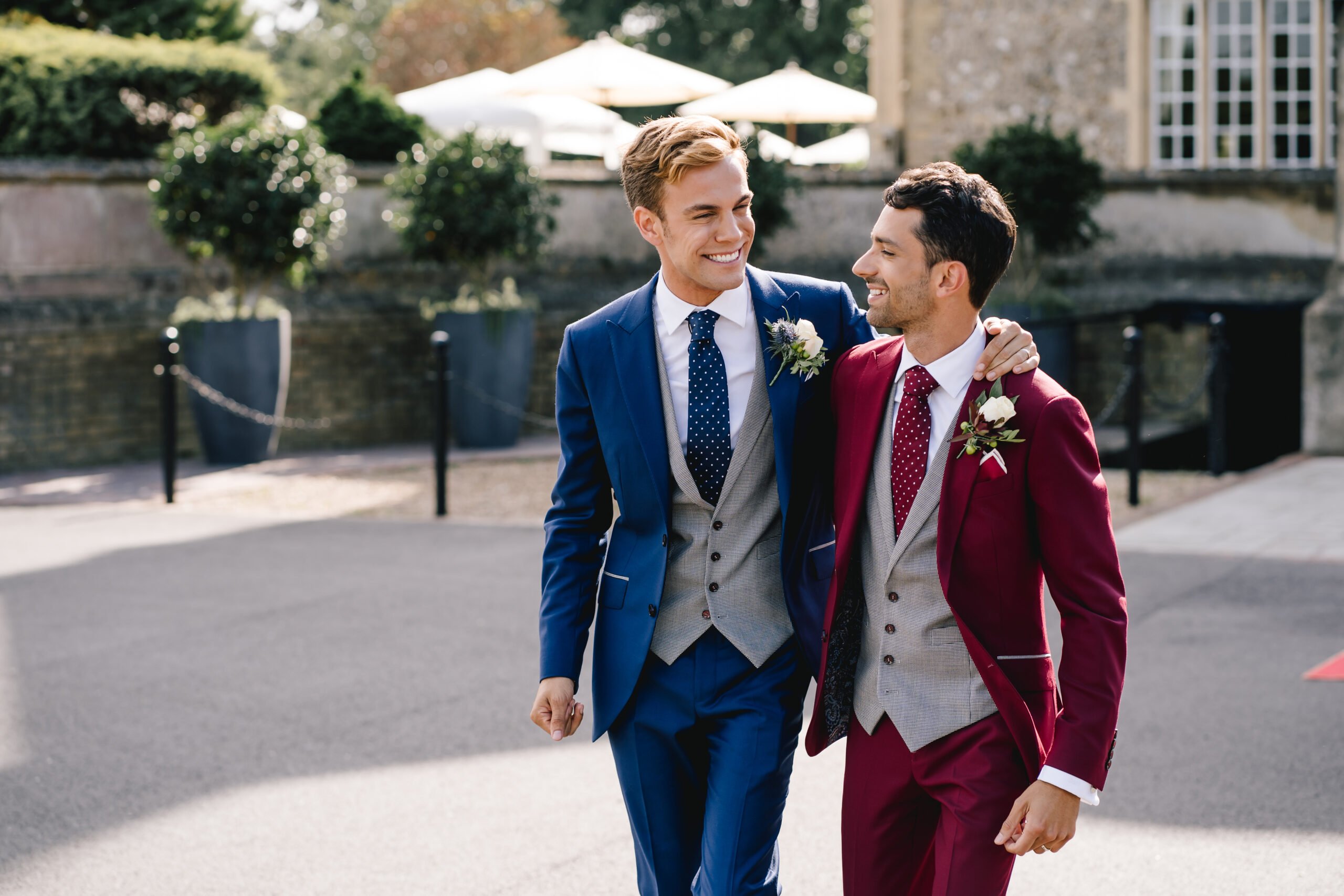 interraciale liefdesfeest van twee mannen in blauw en rode trouwpakken terwijl ze van een schilderachtige trouwlocatie en ceremonieplek vertrekken.