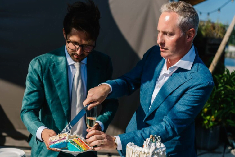 Wedding ceremony readings for gay couples. Two men dressed in green and blue suits are cutting a rainbow wedding cake.