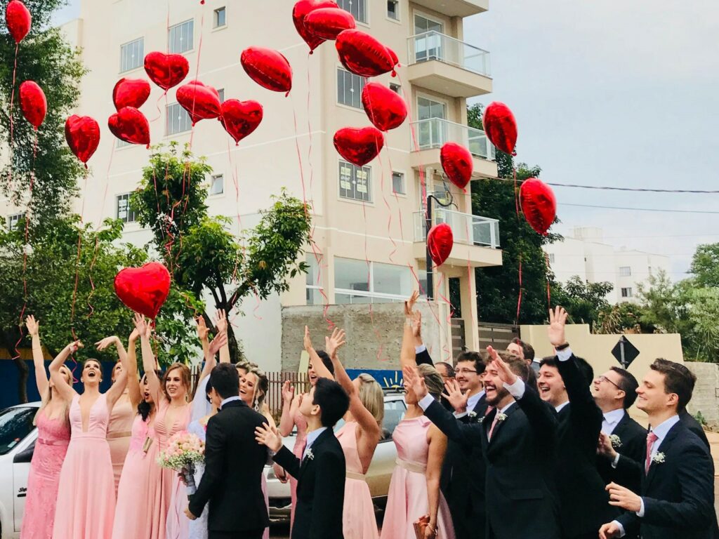 Bride and groom kissing surrounded by bridesmaids dressed in pink and groomsmen dressed in black tie suits holding red balloons.