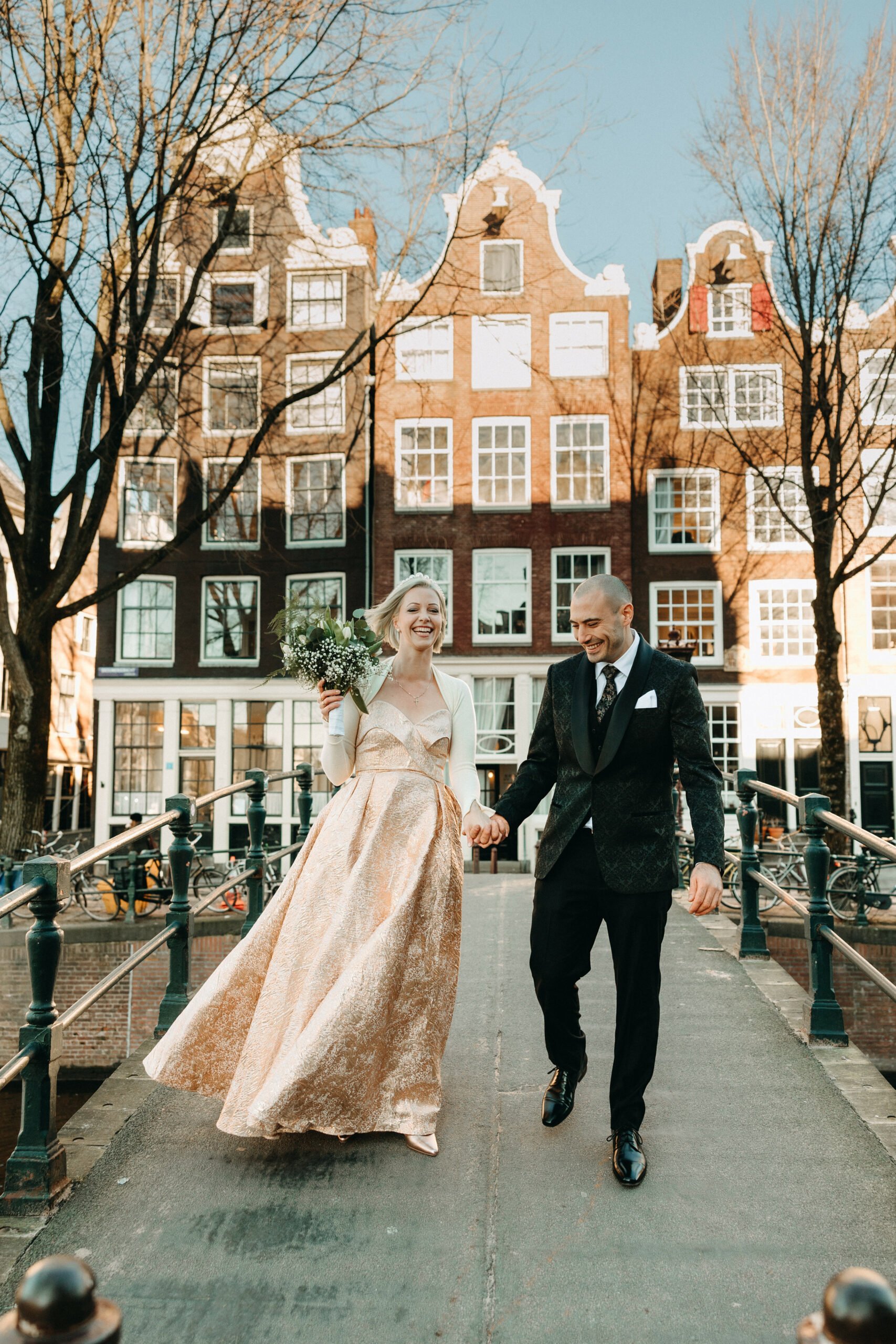 a newly married couple walk across a canal bridge with a backdrop of canal houses of Herengracht, Amsterdam