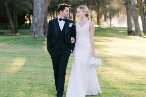 Vondelpark wedding in Amsterdam: bride in white lace dress and groom in black tuxedo and bow tie walking arm in arm in the park on a sunny day.