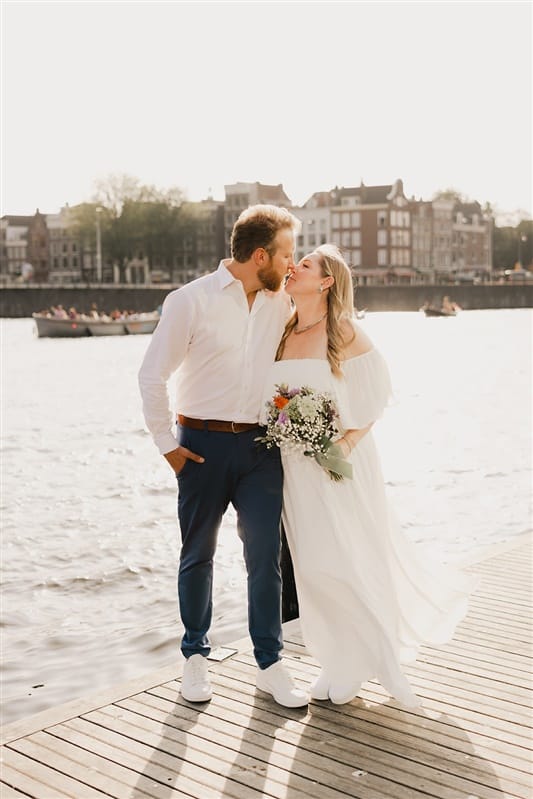 Bride and groom dressed in white holding one another at the canalside in Amsterdam, the Netherlands.