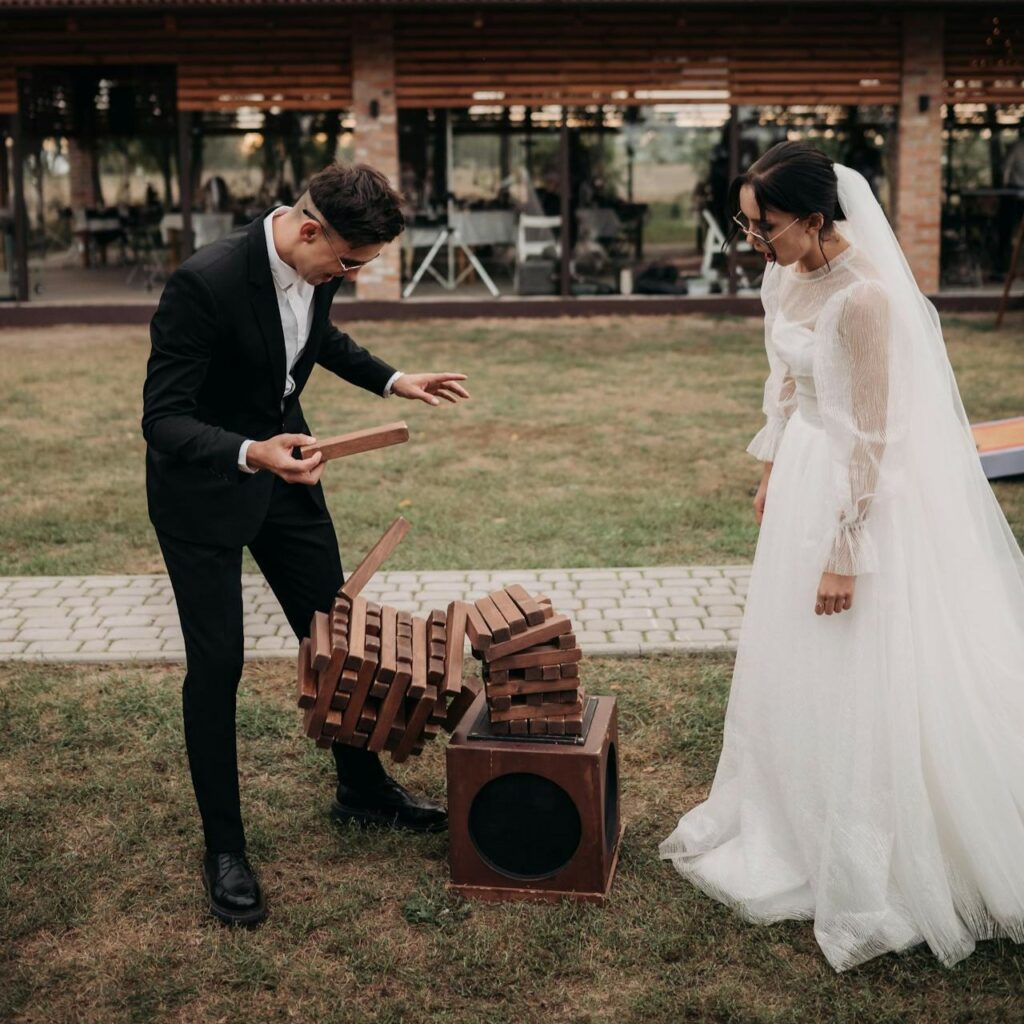 Spiritual wedding rituals, a couple playing a game of Jenga woven into the ceremony. The bride dressed in a white dress is looking at the groom dressed in a black tuxedo holding a Jenga piece. The jenga structure has fallen.