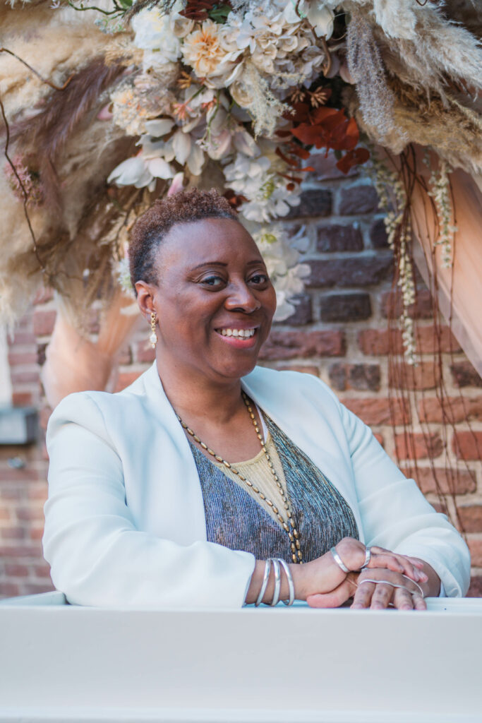 Smiling Black bilingual, international wedding celebrant and ceremony speaker for biracial couples in front of a wedding arch adorned with a background of beige, orange, and flowers. 