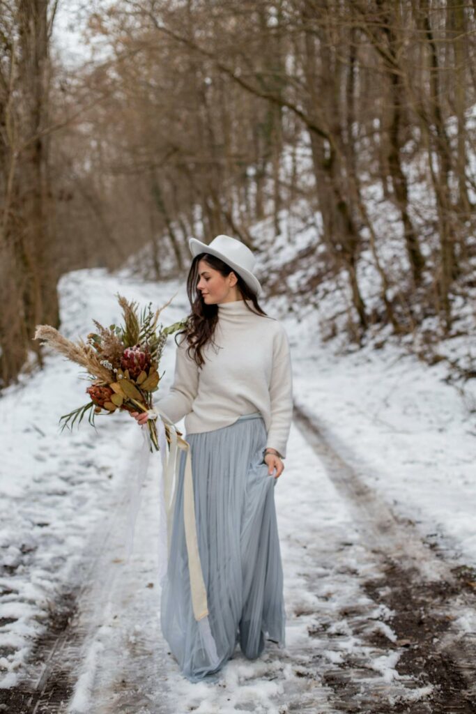New Years' wedding woman-in-a-White-Hat-and-Long-Blue-Long-Skirt-Walking-in-Winter-Forest-with-Dry-Bouquet.jpg